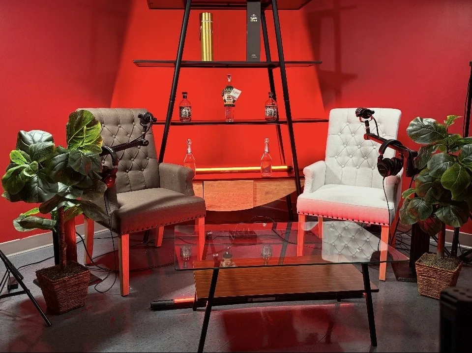 Two armchairs with microphones attached, flanking a glass coffee table, with potted plants on each side, set against a red wall with shelves and bottles.
