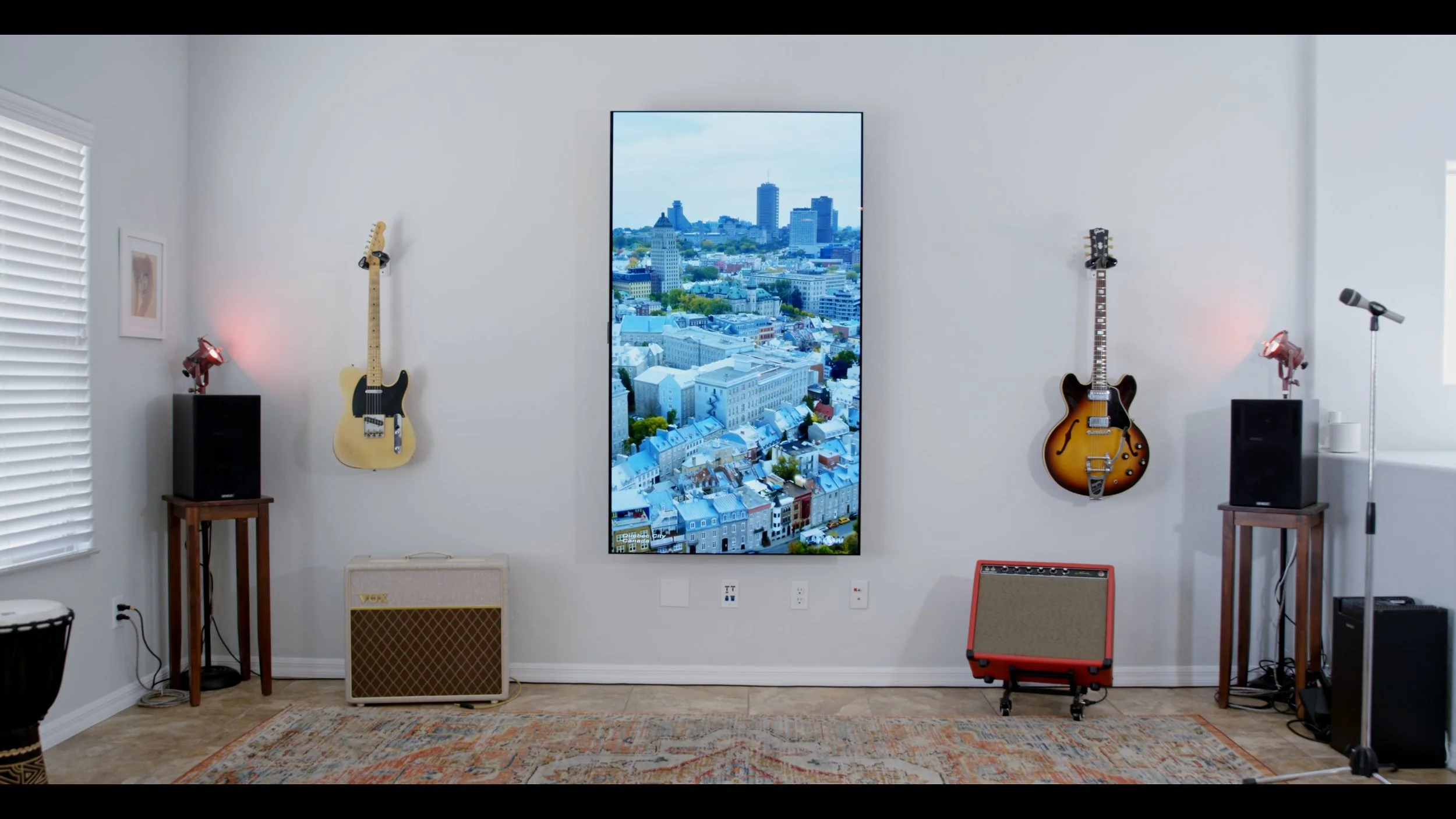A music room with a large vertical cityscape photo or screen on the wall, two electric guitars on the wall, two speakers on stands, a microphone, and a small amplifier on the floor, all on a light-colored hardwood floor with a patterned rug.