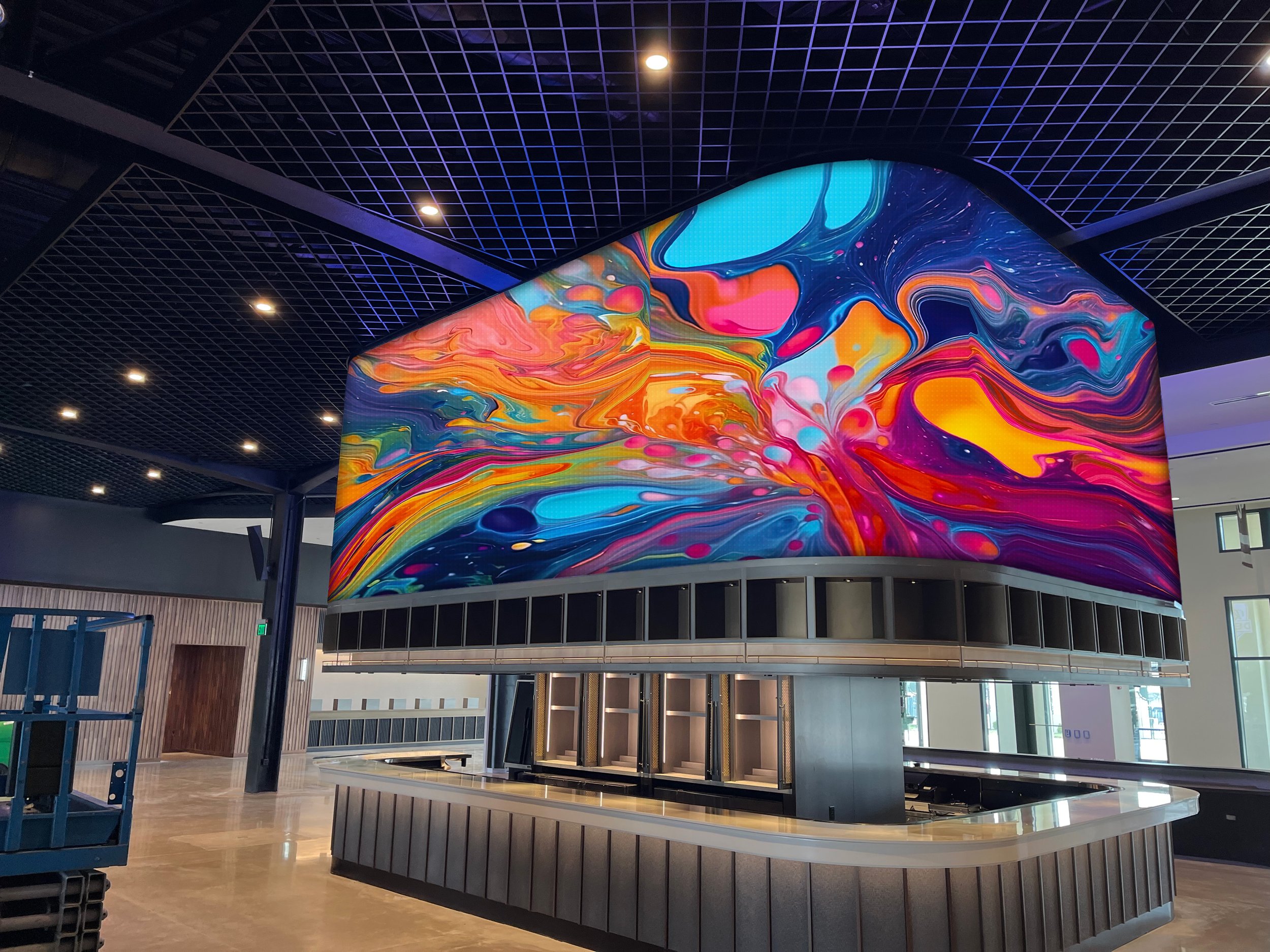 A large, curved LED screen displaying a vibrant, swirling abstract pattern of blues, pinks, and yellows, mounted above a modern bar counter.