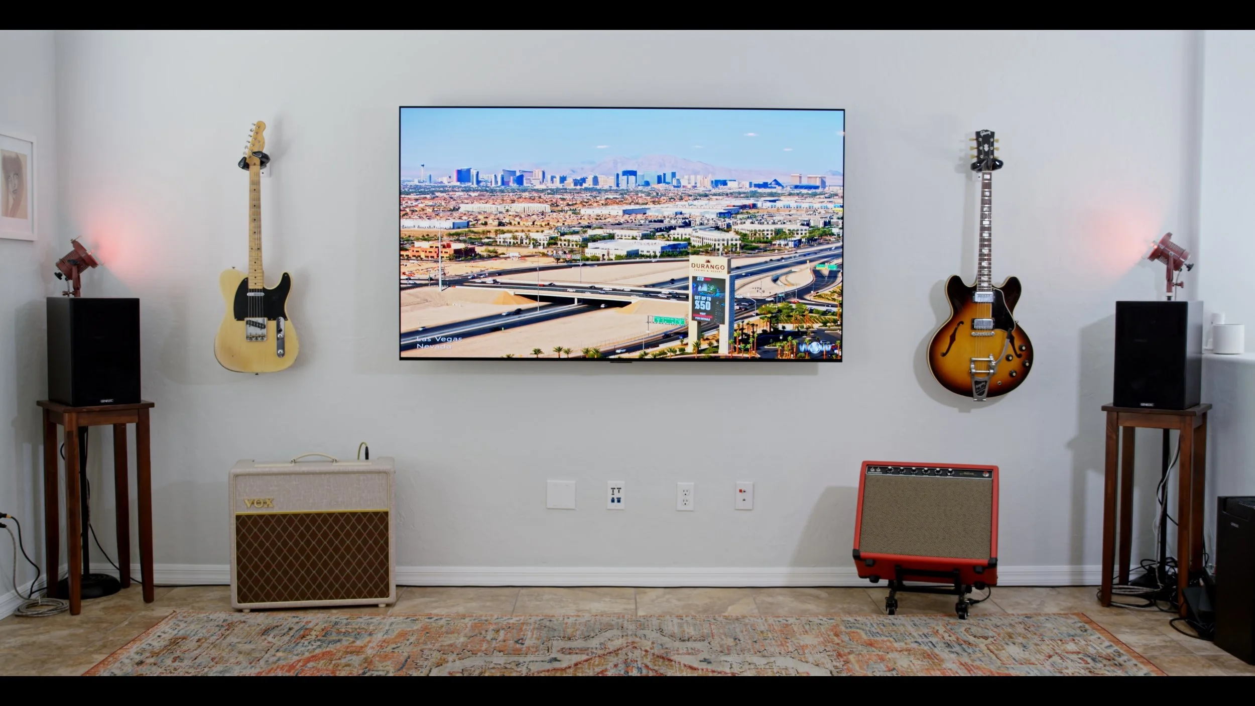 Living room wall with two guitars on either side of a large flat-screen TV, surrounded by speakers, a Vox amplifier, and decorative lighting, on a colorful rug.