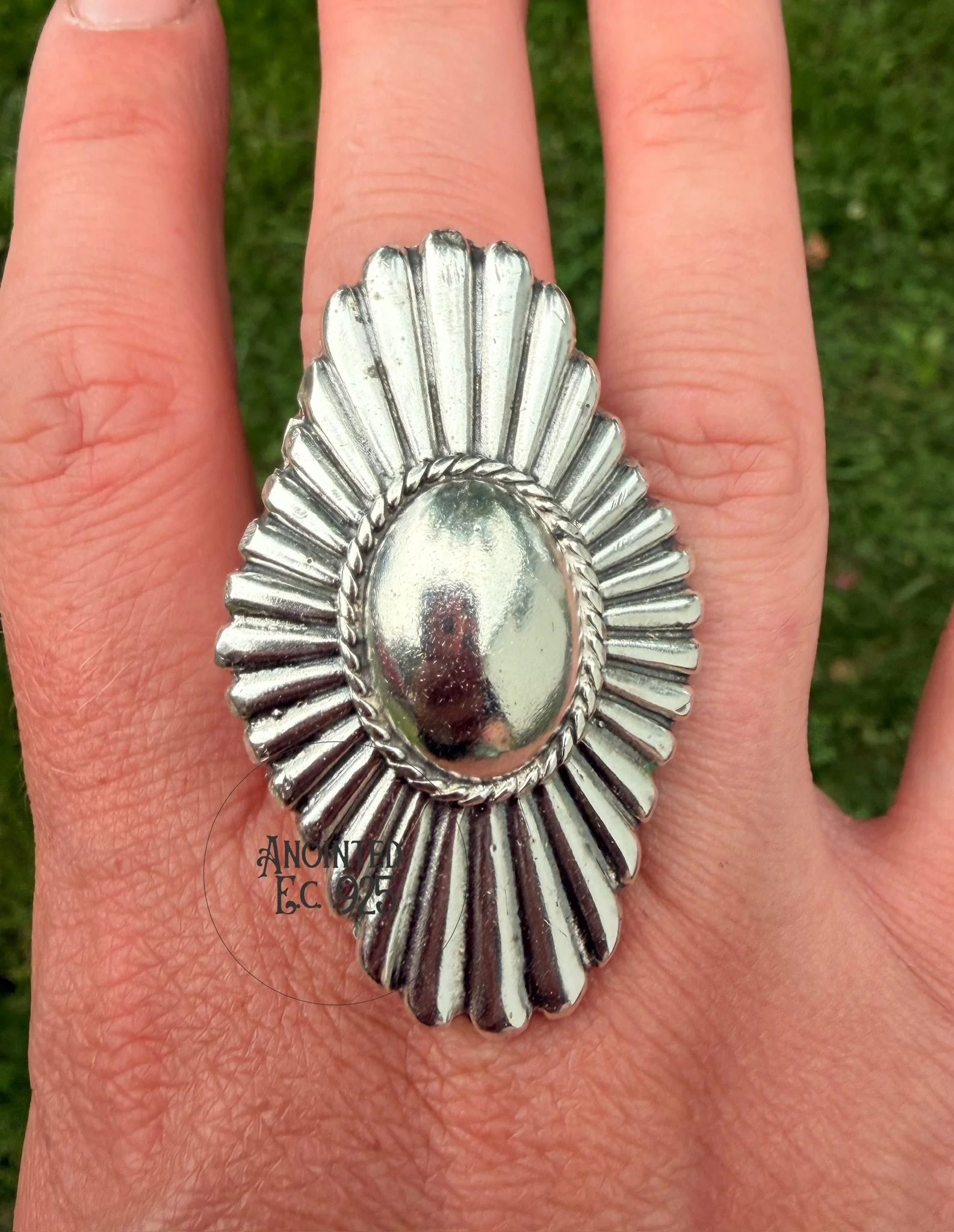 Hand wearing a large silver ring with a shell design, on a background of green grass.