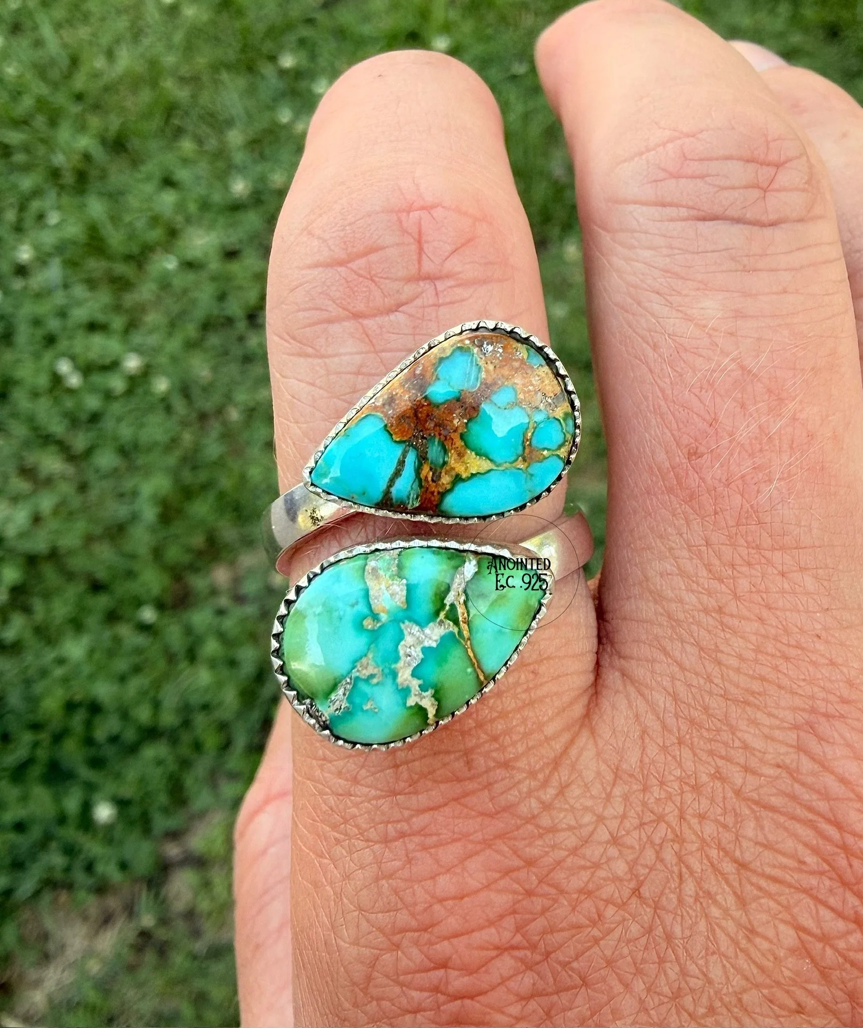 Close-up of a hand wearing a turquoise and multicolored stone ring, with a grassy background.