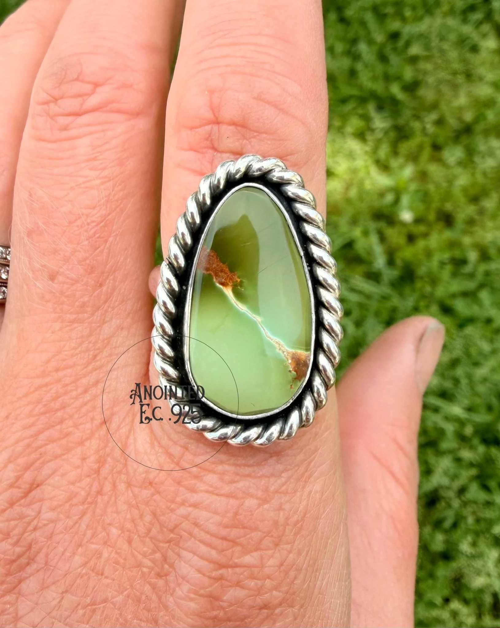 Close-up of a hand wearing a large silver ring with a green stone, against a background of green grass.