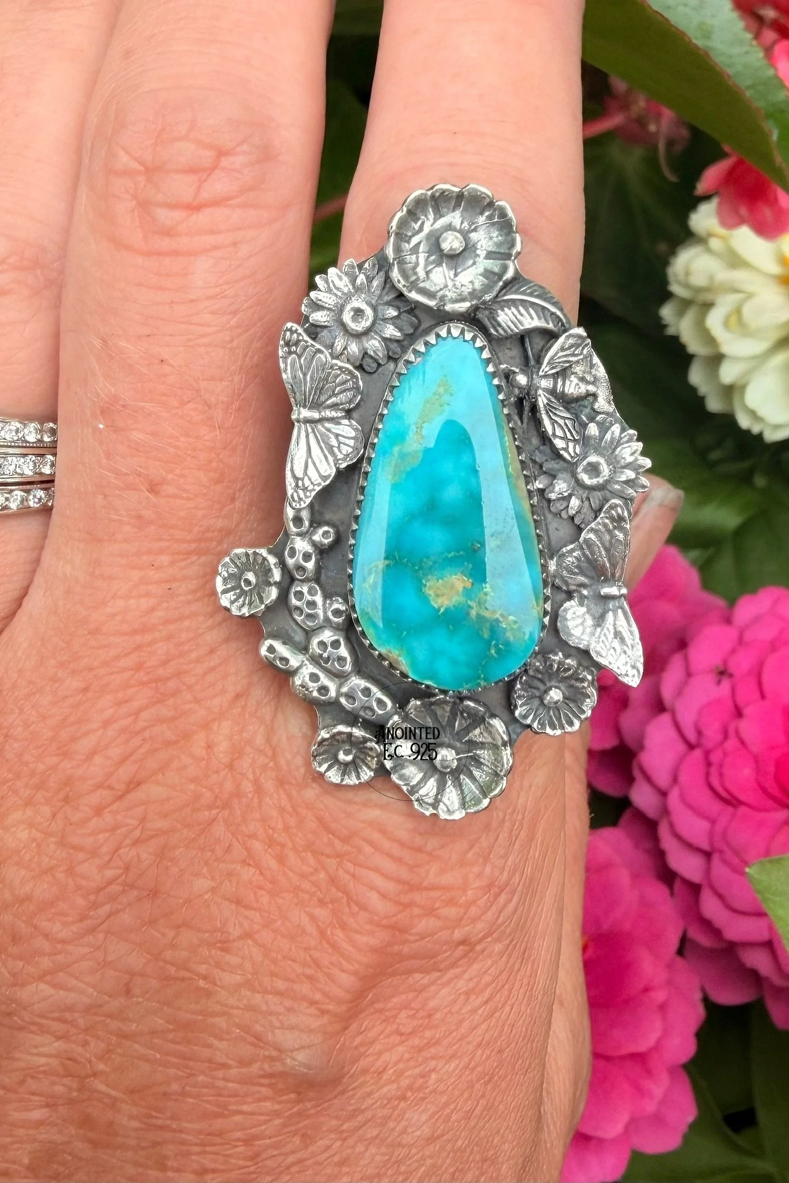 A silver ring with a large turquoise stone, adorned with floral and butterfly designs, worn on a person's finger with a background of pink and white flowers.
