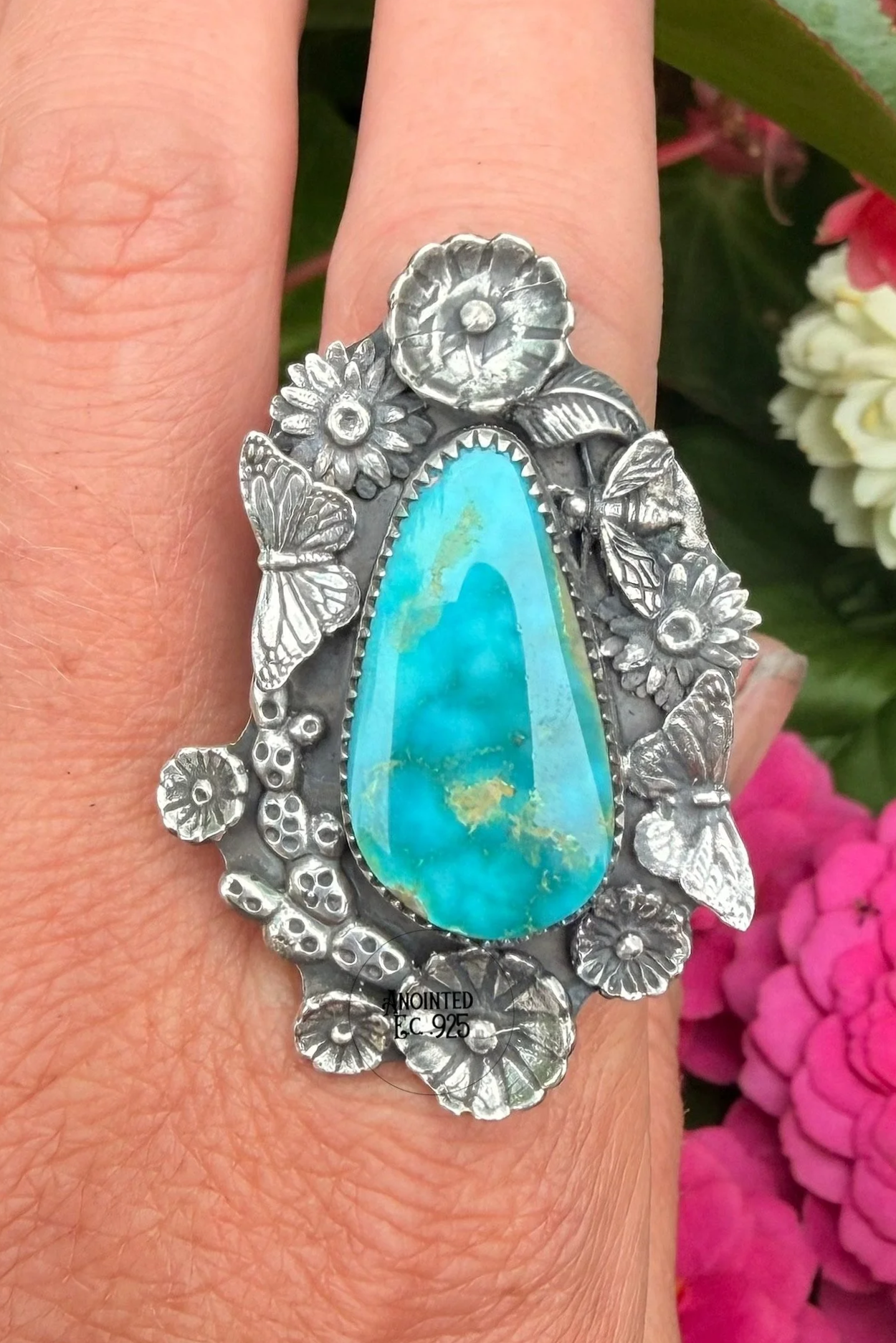 A silver ring with floral and butterfly designs featuring a large, oval turquoise gemstone on a person's middle finger. Background includes pink and white flowers.