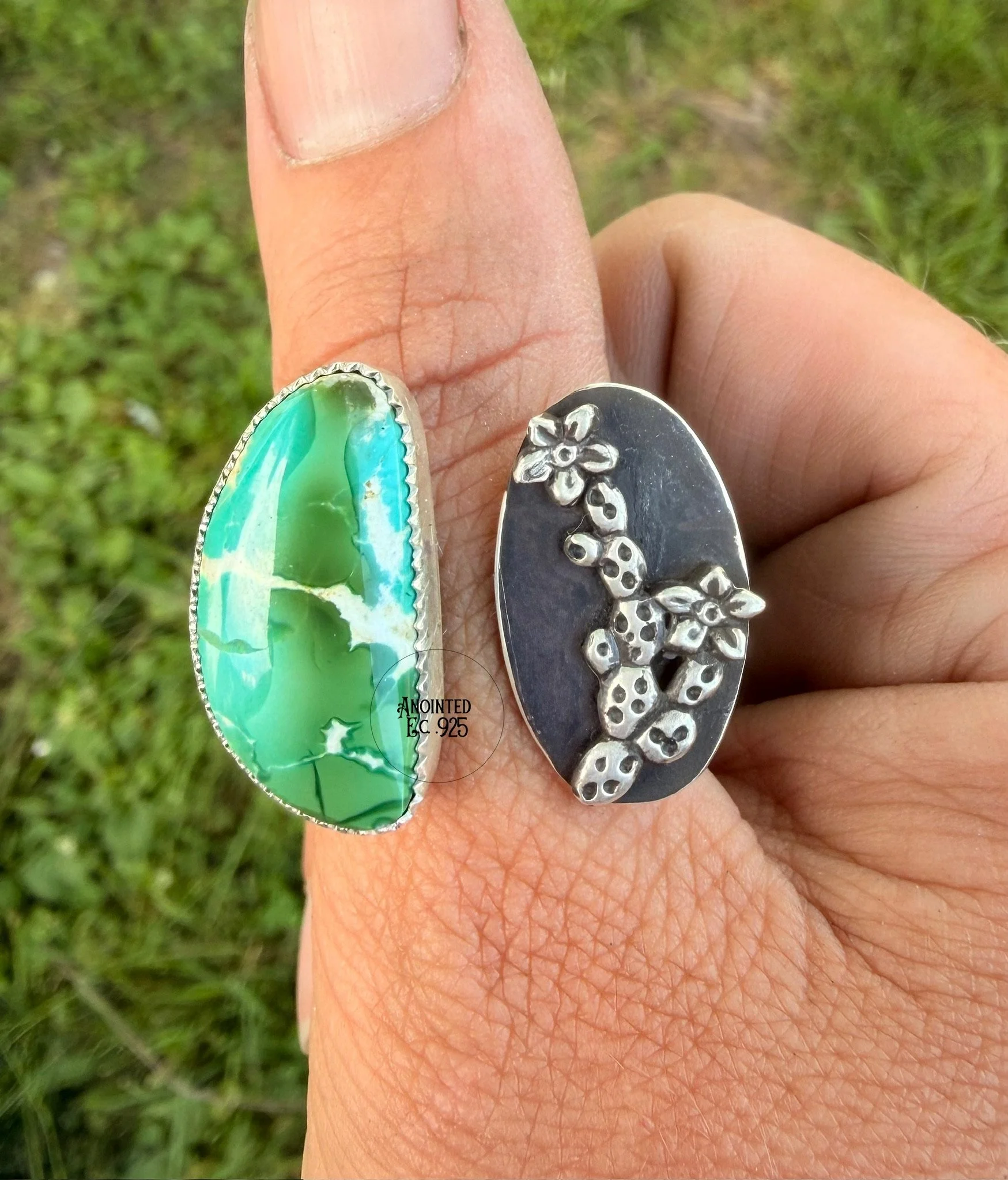 Close-up of a person's hand showing two rings, one with a large oval green and white stone and the other with a floral design on black background, against a grassy outdoor background.
