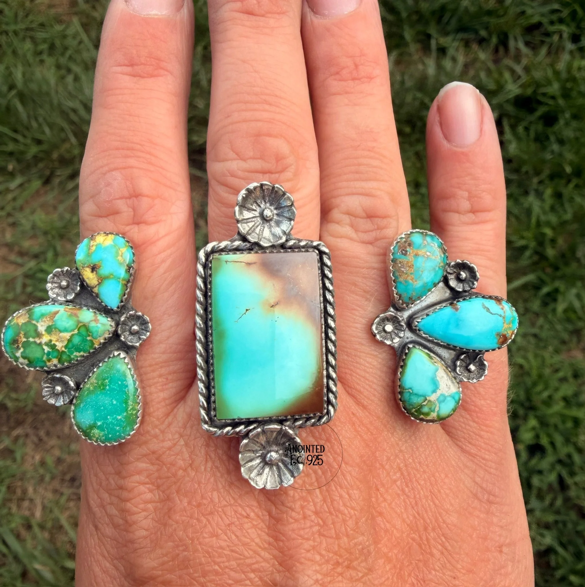 A close-up of a person's hand showing several rings with turquoise stones, including a silver ring with a floral design and a large rectangular turquoise stone with a silver border. The person's index finger is slightly extended. The background is gr