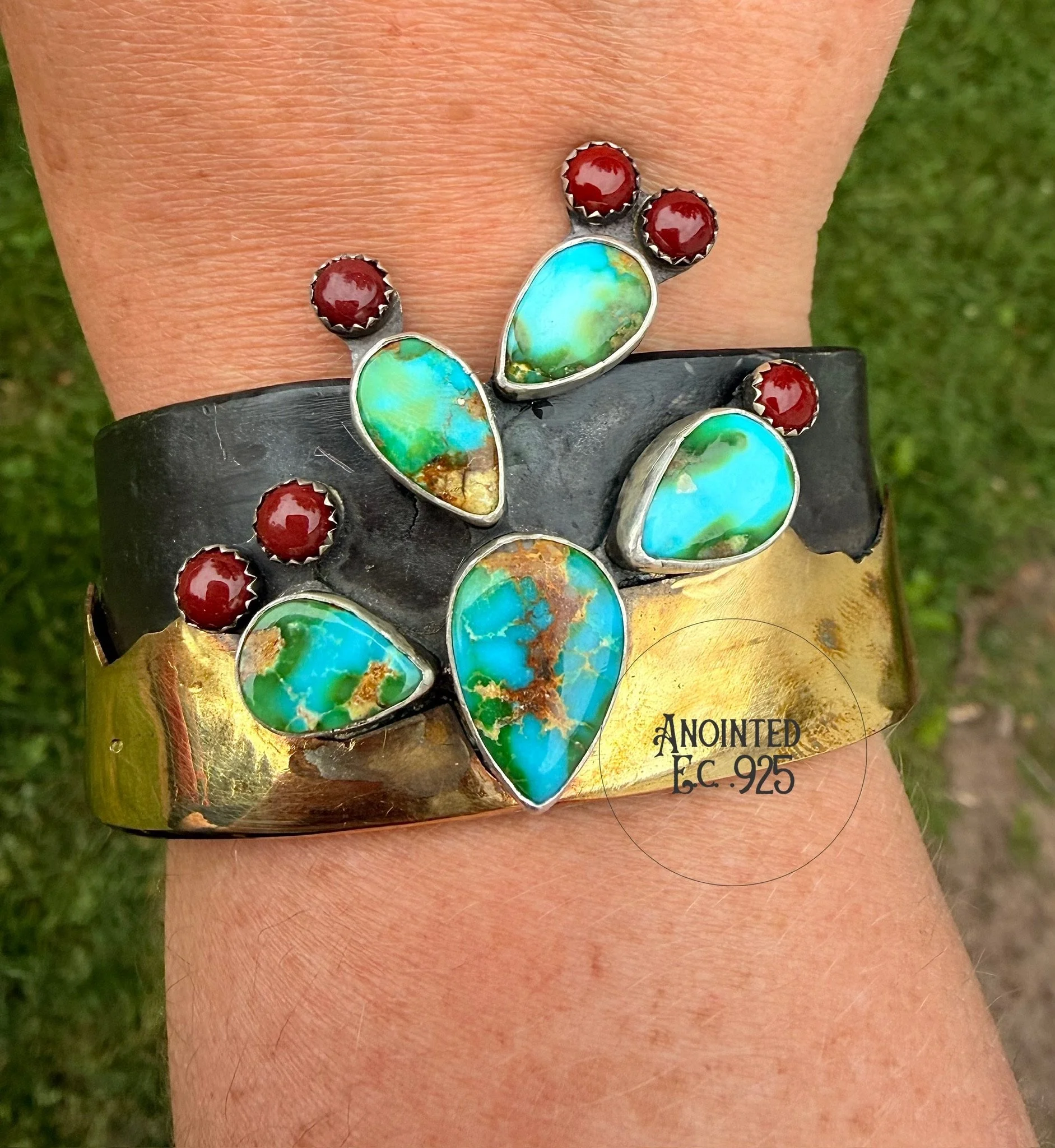 A close-up of a silver ring with large greenish-blue opal gemstones and smaller red gemstones, worn on a person's finger against a blurred green grass background.