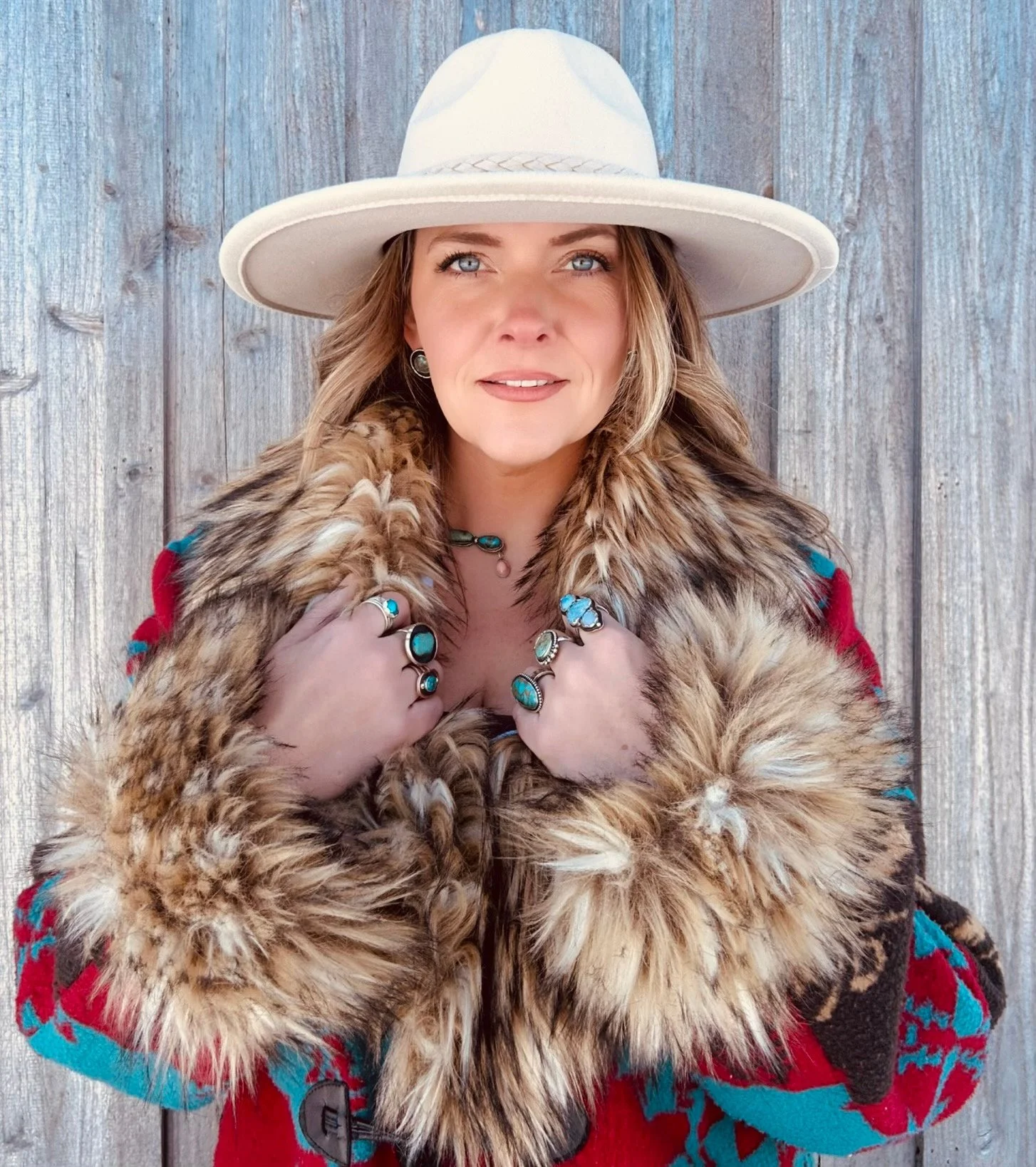 A woman wearing a wide-brimmed white hat and a fur-lined coat, standing in front of a wooden fence.