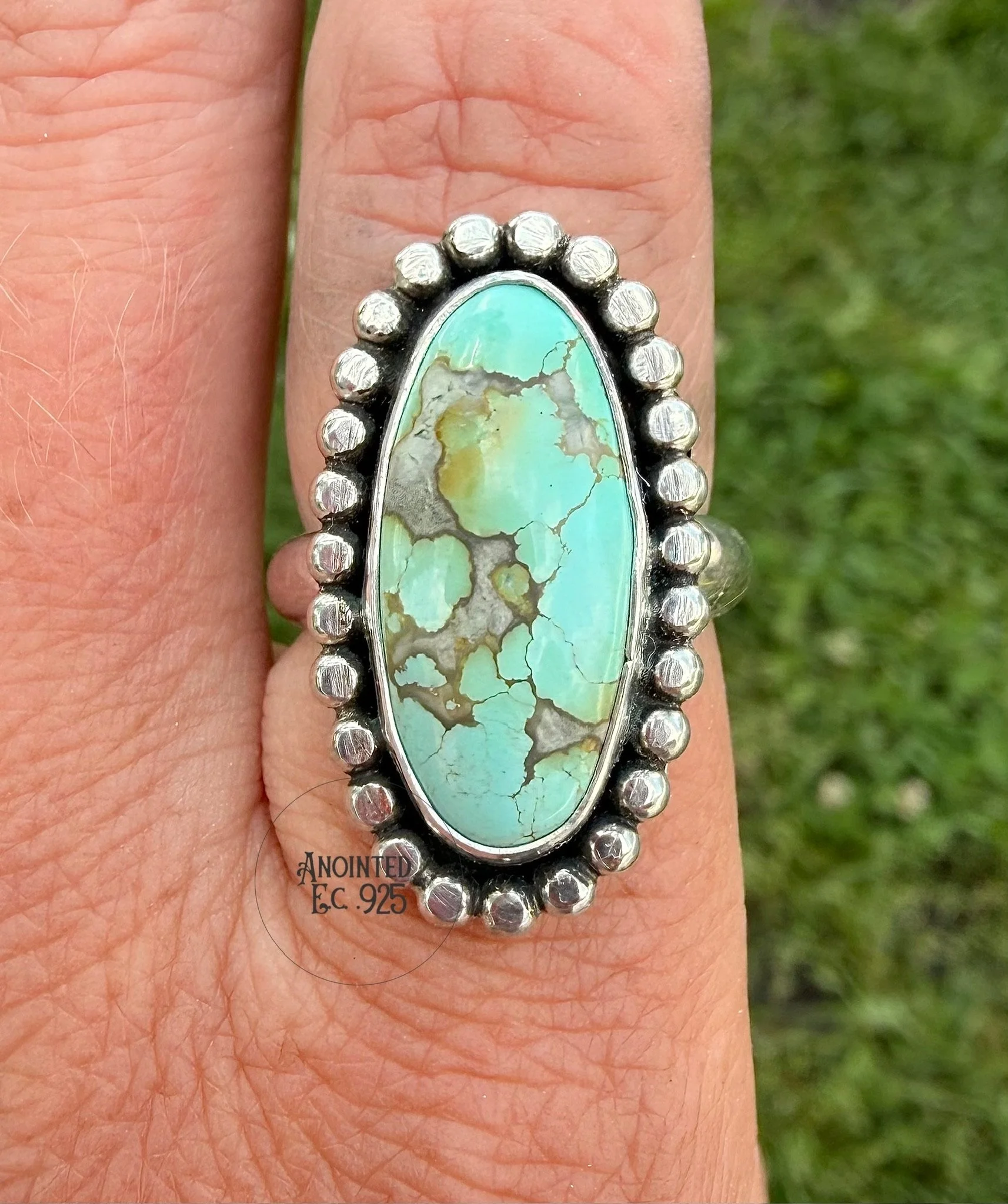 Close-up of a hand wearing a silver ring with an oval turquoise stone with a web of black and light brown matrix. The background is grassy.