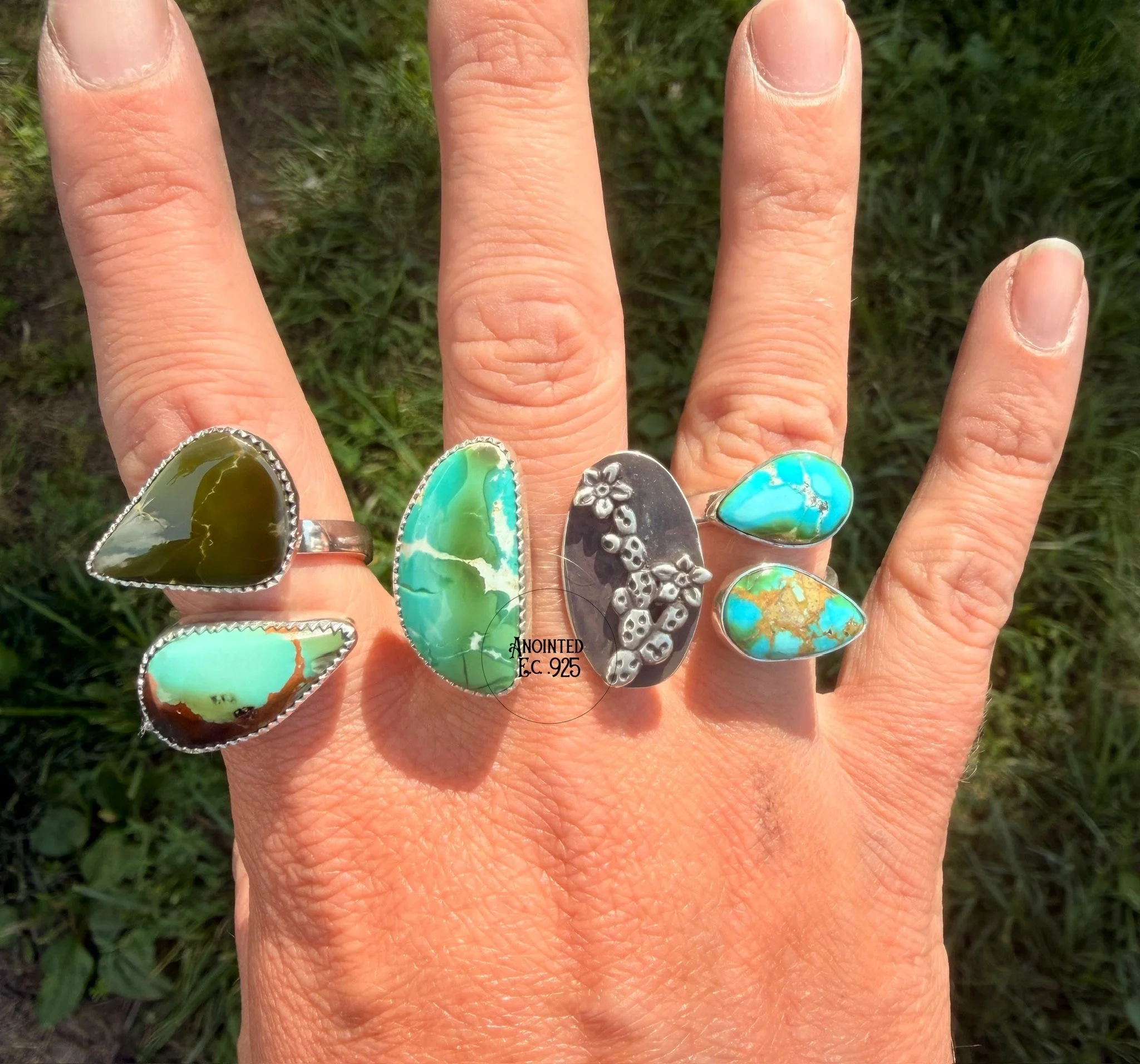 A person's hand with multiple rings featuring turquoise, silver, and green stones, held against a grassy background.