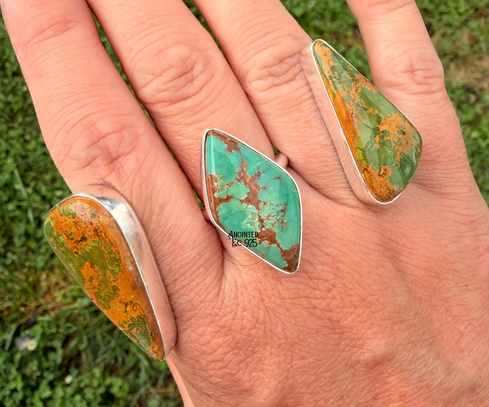 A hand wearing large turquoise and orange patterned gemstone rings against a grassy background.
