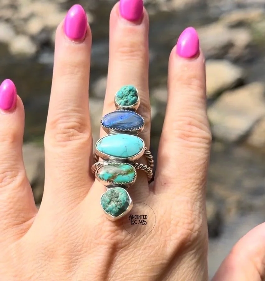A hand with pink nail polish showing five turquoise and blue turquoise gemstone rings, with a rocky outdoor background.