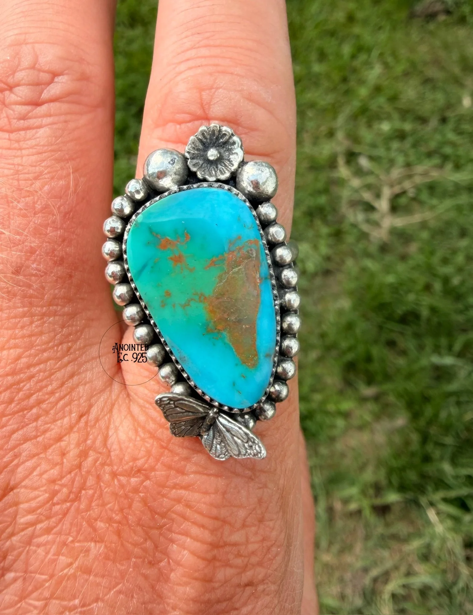 Close-up of a person's hand wearing a silver ring with a large turquoise stone and decorative floral and butterfly designs, set against a grassy background.