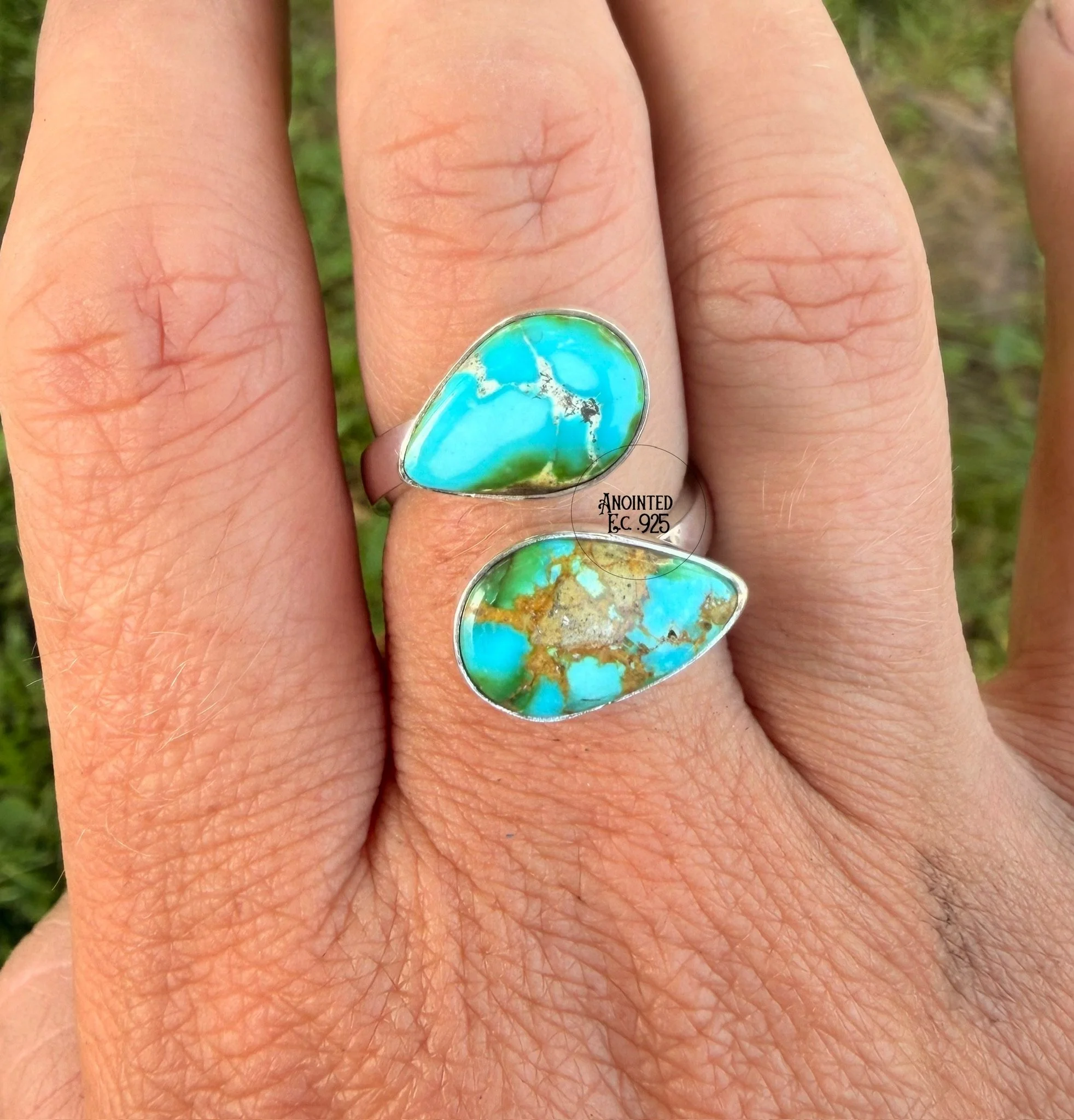 Close-up of a hand with three fingers visible, each wearing rings with turquoise stones. The background is blurred green foliage.