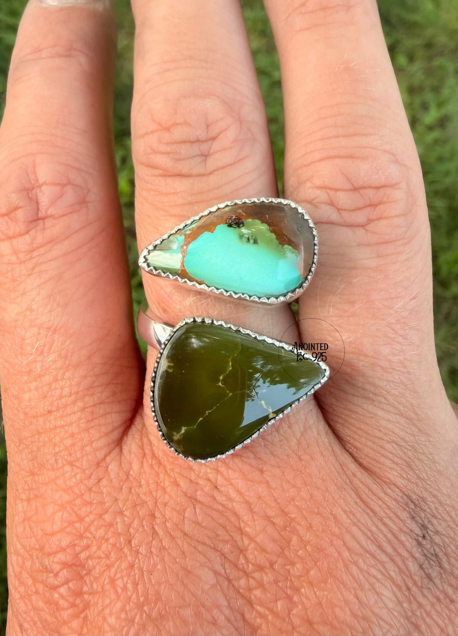 Close-up of a hand with three large rings, each with different colored stones, on the fingers, with a background of grass.