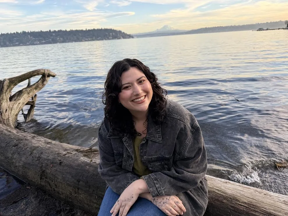 A woman with dark curly hair smiling, sitting on a fallen tree by a large body of water, with trees and a distant shoreline in the background during a sunset.