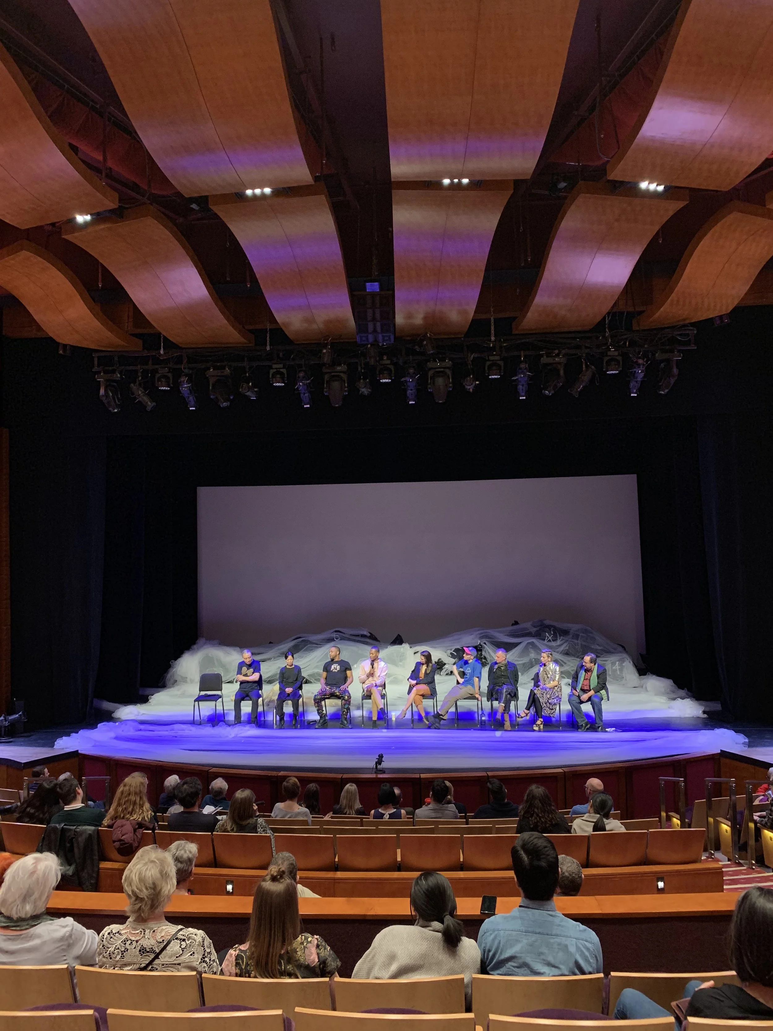 Image of a theatre stage with a row of performers, the seats in front of the stage are also evidence