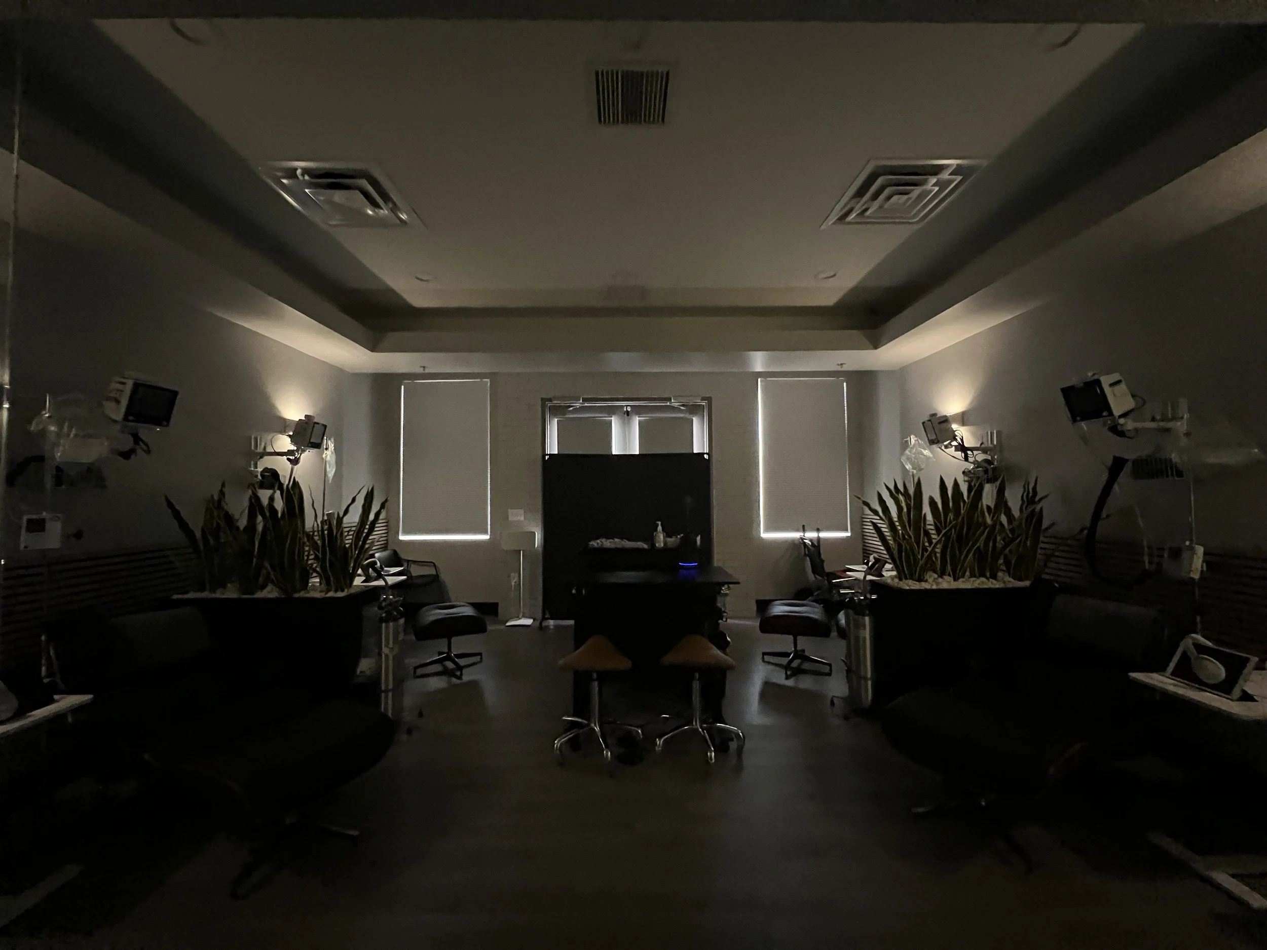 Dimly lit hospital or medical room with two windows, seating, plants, medical equipment, and monitors on the walls.