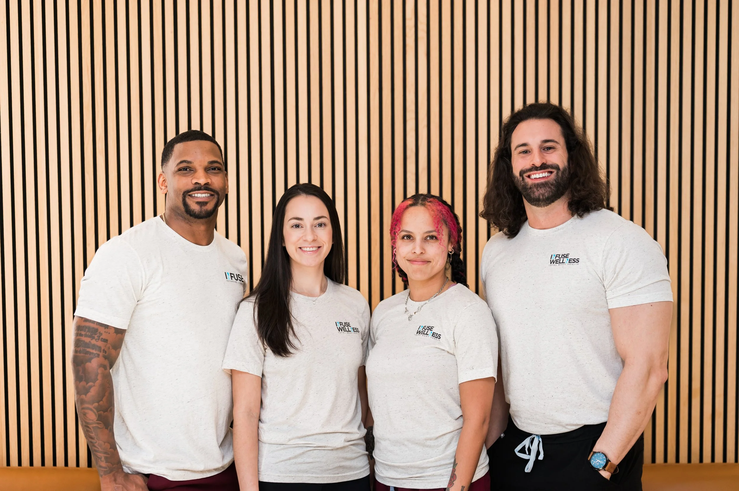 Four diverse people standing in front of a wooden slat wall, smiling, wearing matching light gray T-shirts with a logo that says 'INFUSE WELLNESS'.