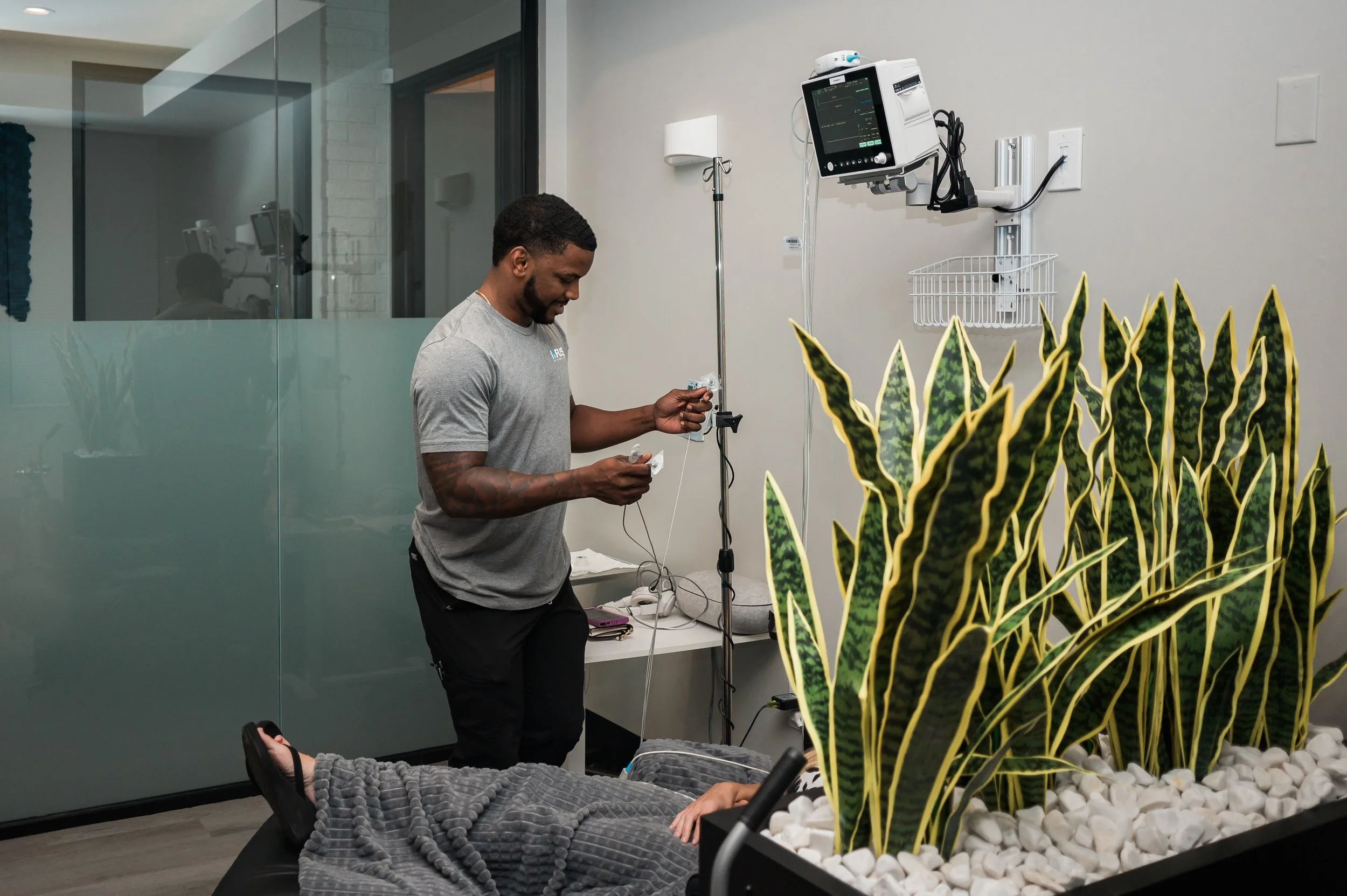 A healthcare worker standing next to a patient lying on a bed in a hospital room, with a large plant in the foreground and medical equipment attached to the patient.