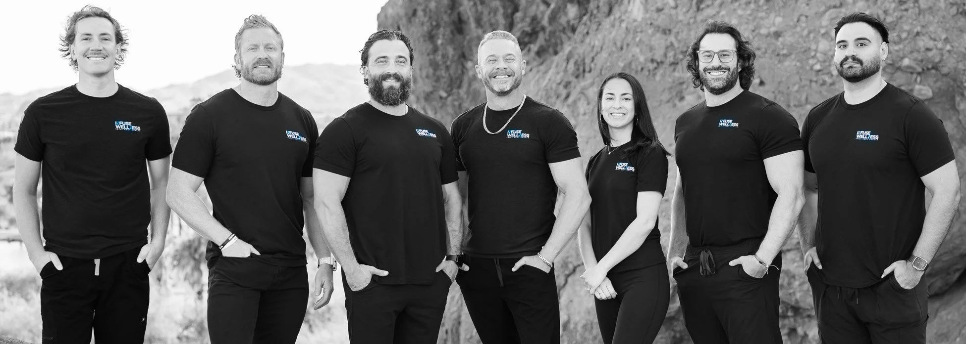Group of seven diverse young adults standing outdoors, smiling, wearing matching black t-shirts with blue and white logo that reads 'Fuse Wellness,' on a mountainous landscape background.