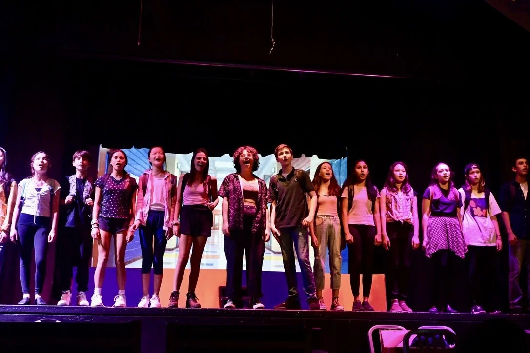 Group of young performers on stage during a theatrical production, with a colorful background projection.
