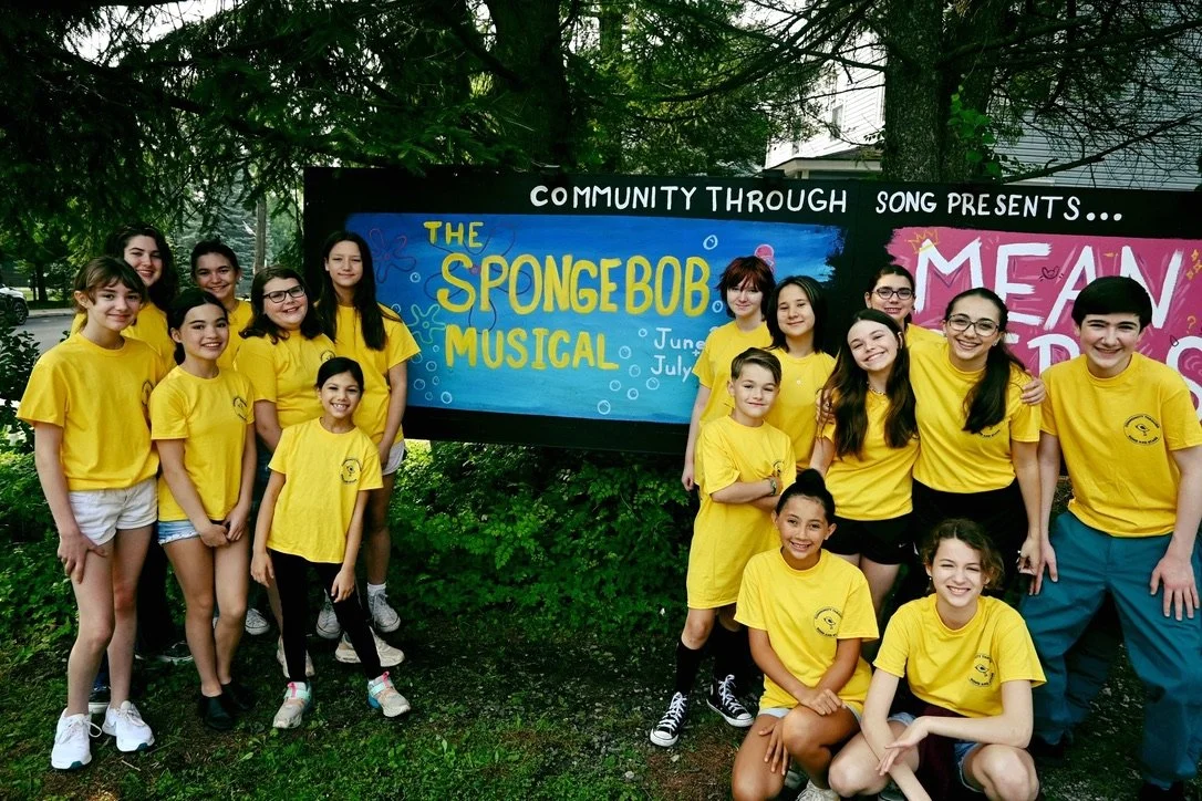 Group of children and teenagers wearing yellow shirts standing outdoors in front of a sign that reads 'The SpongeBob Musical'.