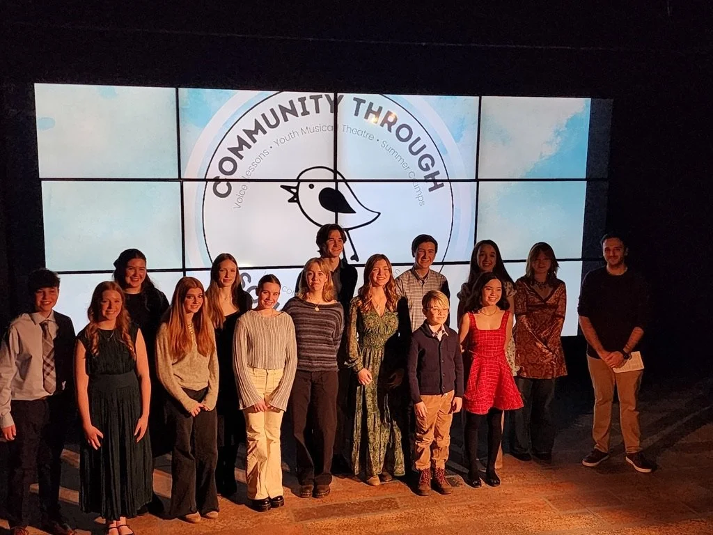 Group of children and adults posing on stage in front of a large screen with the logo of Community Through, a youth musical theater summer camp, featuring a bird and blue sky background.