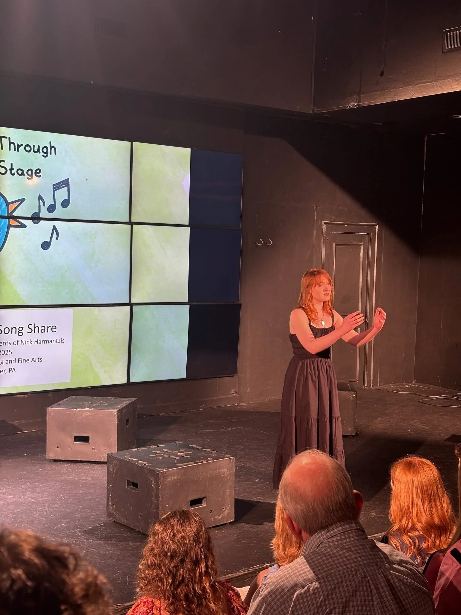 A woman is performing or speaking on stage in front of an audience. There is a large screen behind her with a presentation slide showing a colorful graphic and text about a song. The audience is seated, watching her.
