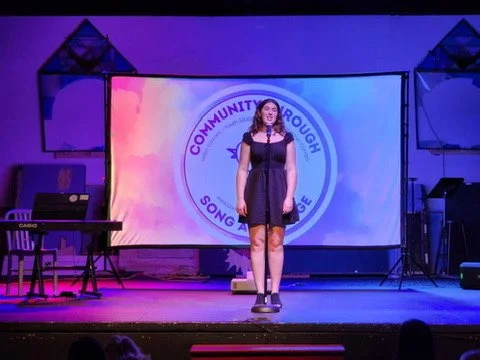 A young woman standing on a stage in front of a large screen displaying a circular logo with the text 'Community Through Song and Speech.' The stage is equipped with musical instruments, including a keyboard and a guitar, under purple and blue lighti