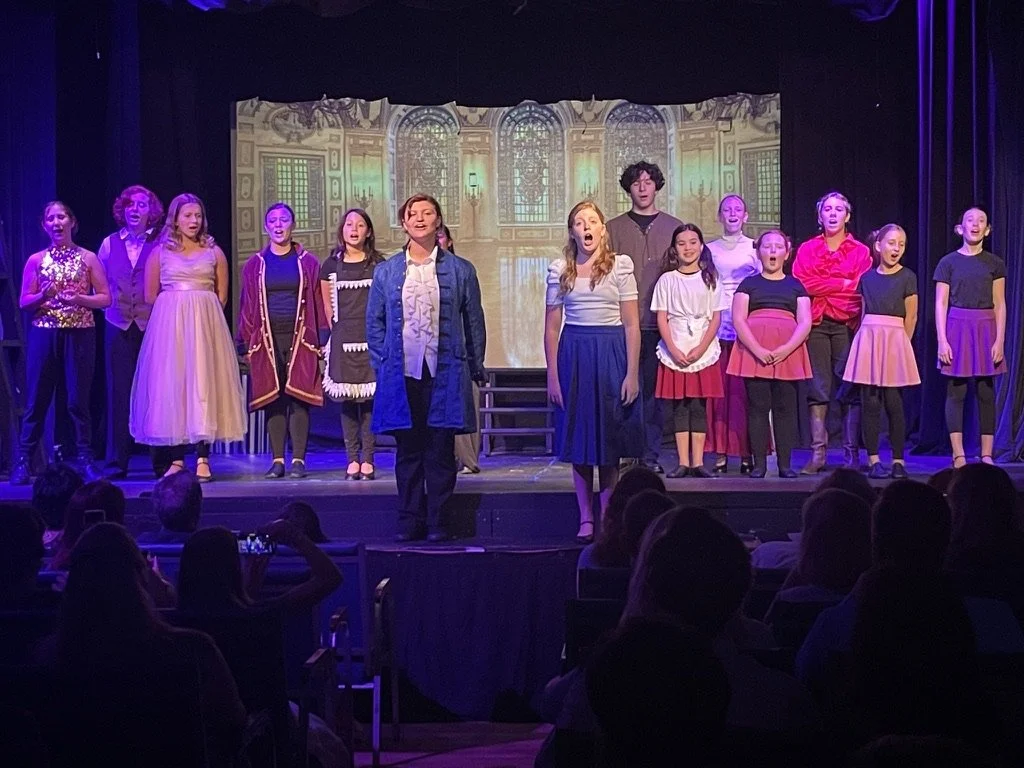 Children performing on stage in a theatrical play, with a backdrop of a mansion or grand building, some children singing or acting, audience visible at the front.