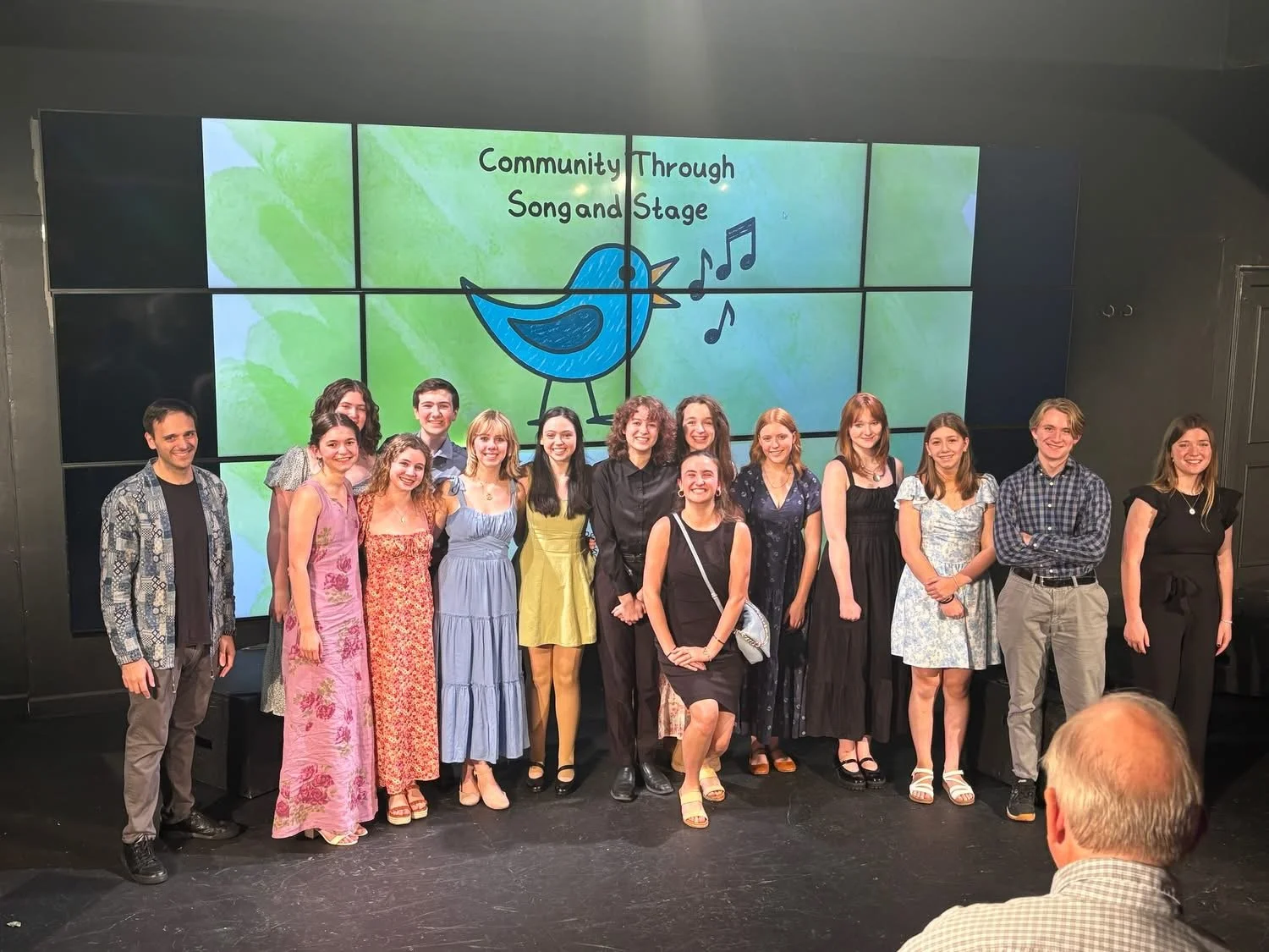 Group of people posing on stage in front of a large screen with a blue bird logo and the text 'Community Song and Stage' at a performance or event.