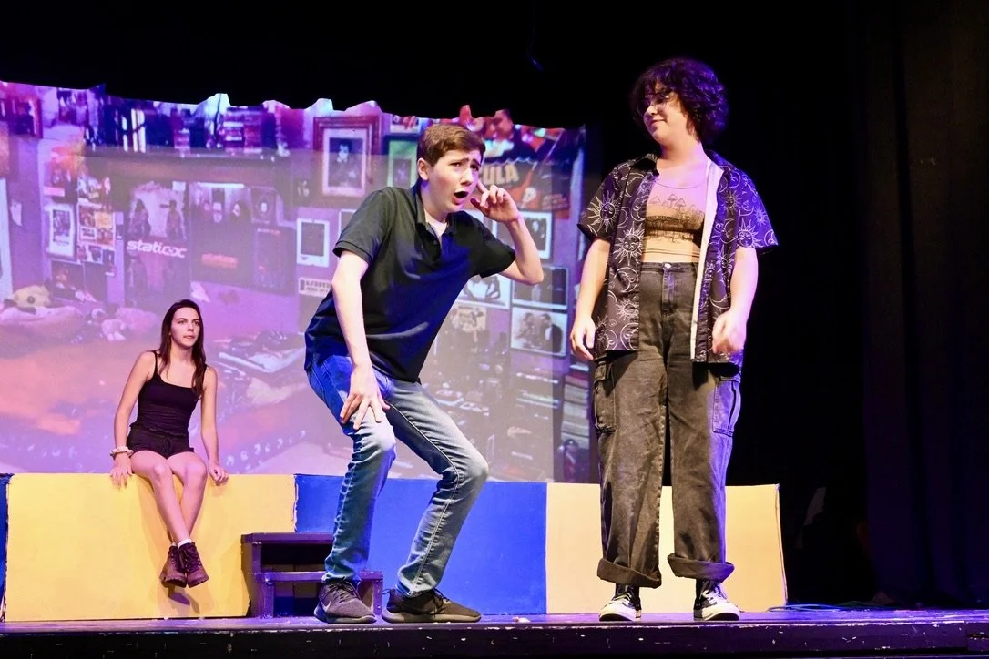 Three young actors performing a scene on stage with a background projection of a cluttered room. One actor is sitting on a yellow block, another is standing and making a gesture, and the third is standing with curly hair and glasses.