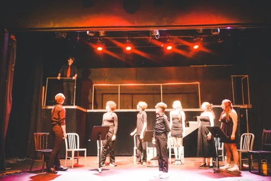 Theater scene with seven actors standing on stage in front of a row of chairs, some with scripts, under stage lighting, with a director or instructor standing on a raised platform observing.