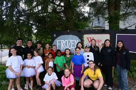 Group of children and teenagers standing outdoors in front of a sign that reads 'FROZEN' and other colorful signs, some wearing costumes and casual clothes.