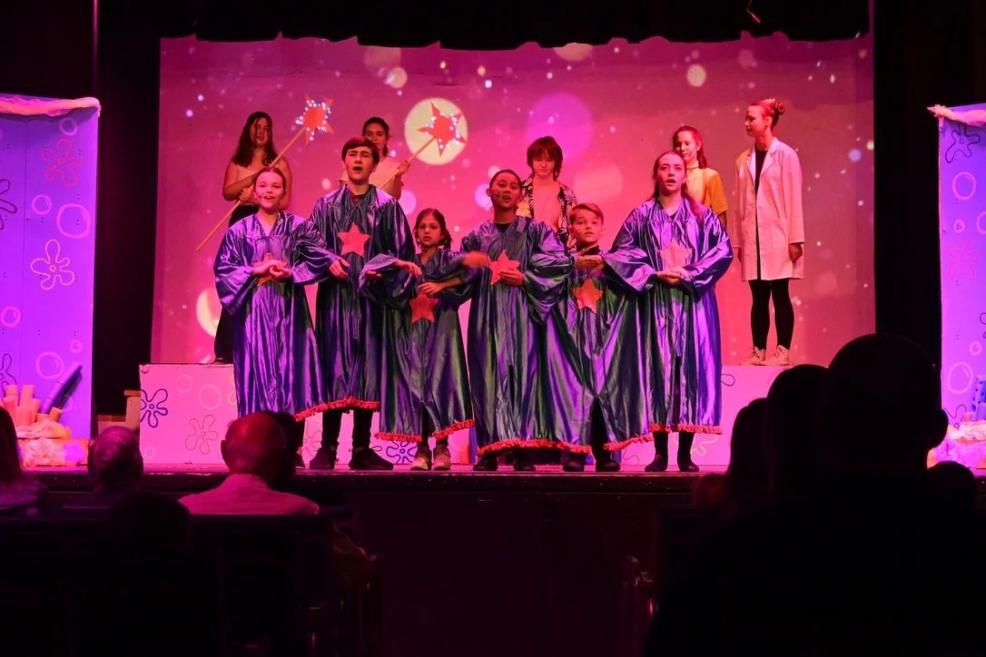 Children on stage performing in a play with a pink background, some dressed in purple robes with star decorations, others in casual clothing, with audience seated in front.