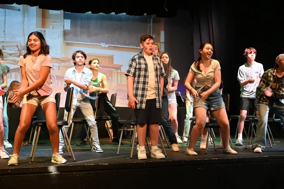 Children performing on stage, some standing and some seated, during a school play or rehearsal with a classroom background projected on the screen behind them.