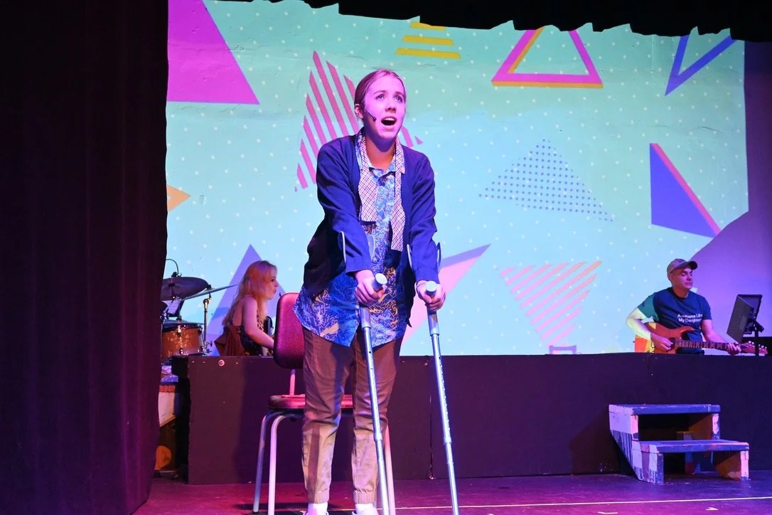 A young woman with crutches standing on stage during a performance, with a colorful geometric background projected behind her, and musicians playing instruments in the background.
