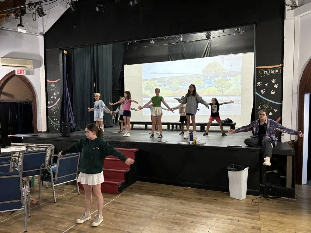 Children rehearsing or practicing on a stage with an adult supervisor, in a room with a wooden floor and theater theater-style seating.