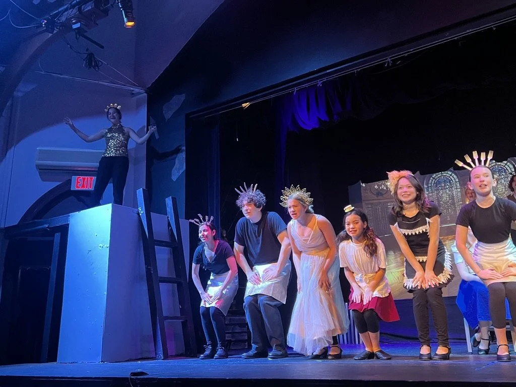 Children performing on stage in a play, wearing costumes with crowns and various outfits, with a person standing at a higher platform observing them.