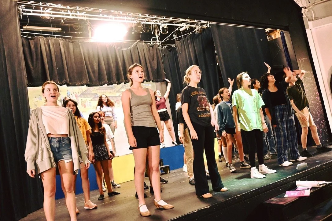 Group of young performers rehearsing on a stage with black curtains and colorful set pieces in the background.