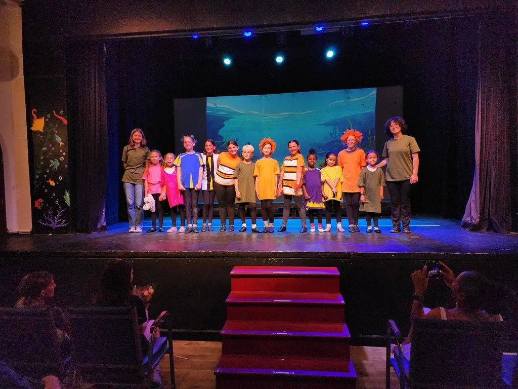 Children and two adults standing on stage after a performance in a theater, with a colorful underwater scene projected in the background.