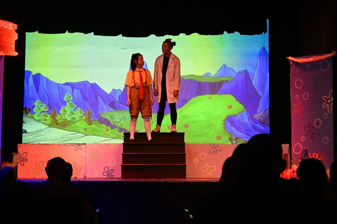 Two children, one in a Scout uniform and the other in a white coat, stand on steps on a stage with a colorful, animated mountain landscape backdrop.