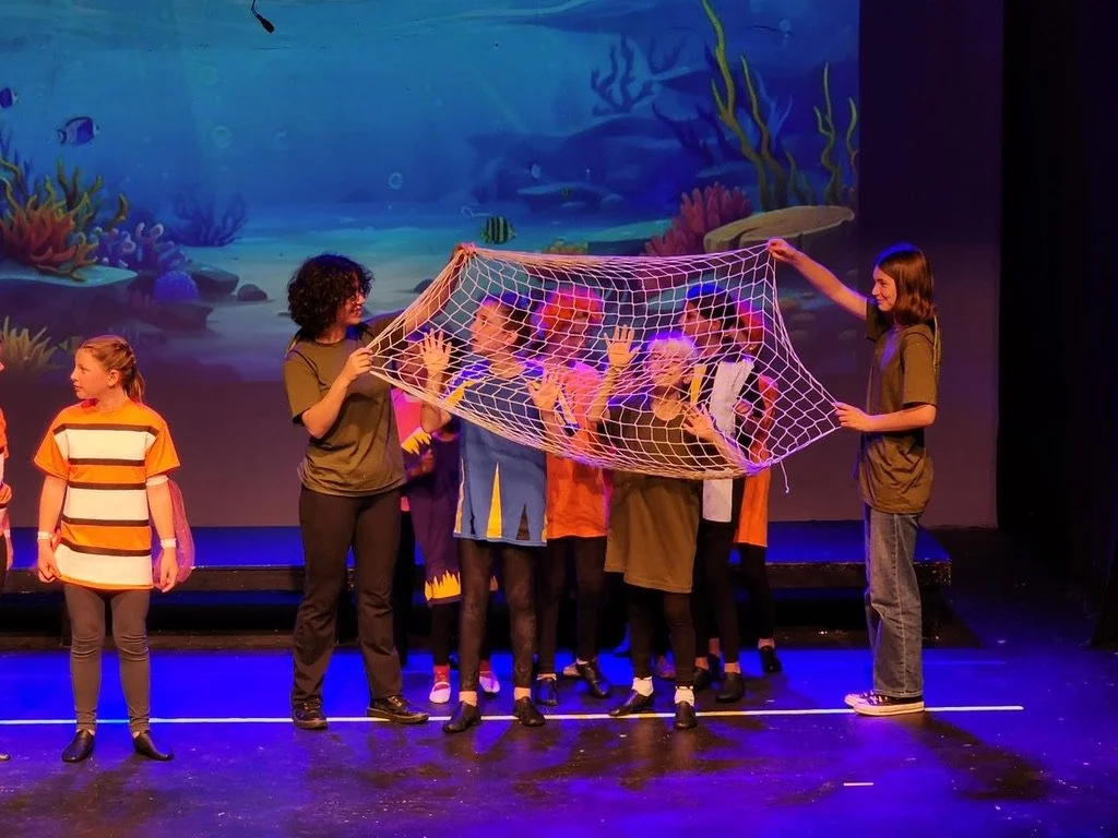 Children performing a play on stage with underwater scene background, holding a net as part of the scene.