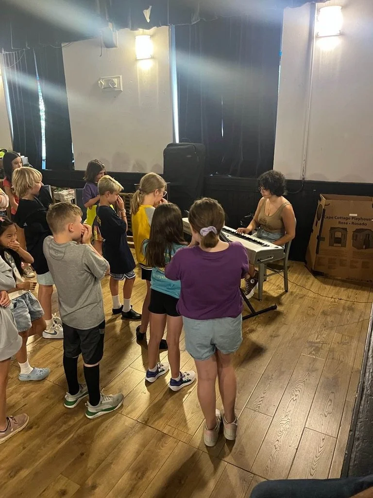 A woman playing a keyboard for a group of children in an indoor setting, with some children standing and others sitting, watching attentively.