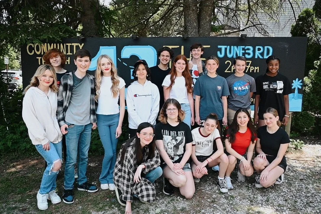 Group of 15 young people posing outdoors in front of a blackboard sign with event details.