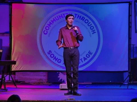Young man in a red shirt and dark pants speaking into a microphone on stage, with a circular logo in the background that reads 'Communicating Through Song Stage'.