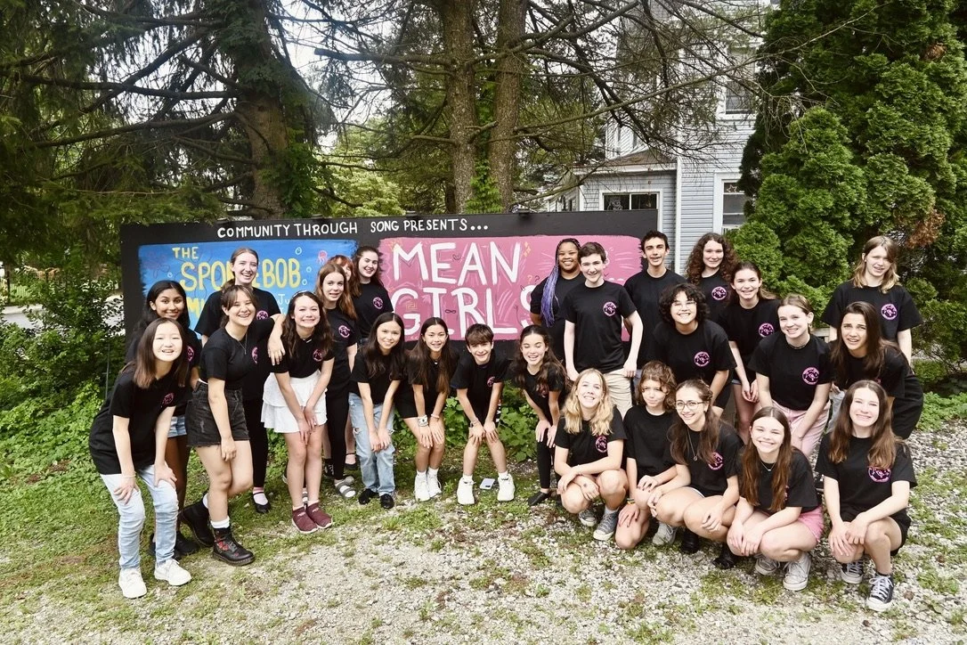 Group of young girls and boys posing outdoors in front of colorful signs that read 'The Sporty Bob' and 'Mean Girls,' with trees and a house in the background.