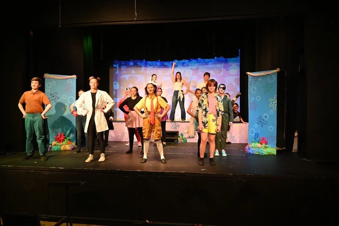 Group of children performing on a colorful stage, some wearing costumes, with a backdrop depicting a cartoon underwater scene.